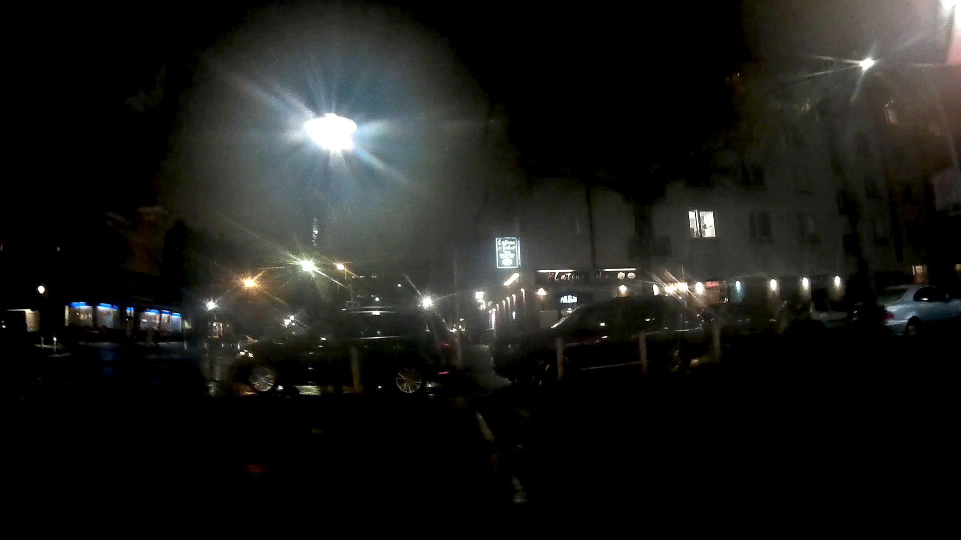 ein bei Dunkelheit mit einer am Fahrrad montierten Dashcam aufgenommenes Bild. Es ist mittig unten eine weisse, gestrichelte Linie zu erkennen die zur Bildmitte hin auf zwei Autos zu läuft, die quer zu einen Fuss- und Radweg abgestellt sind, so das nur eine Lücke bleibt durch die sich ein Fussgänger zwängen kann.