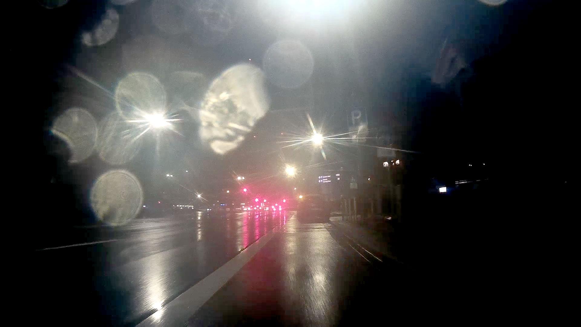 ein mit einer an ein Fahrrad montierten Dashcam bei Dunkelheit aufgenommenes Foto. Da Fahrrad bewegt sich auf einem mit der Autospur mitlaufenden Fahrradweg, der durch einen weissen Streifen abgetrennt ist. Der Radweg wird komplett von einem geparkten Auto blockiert