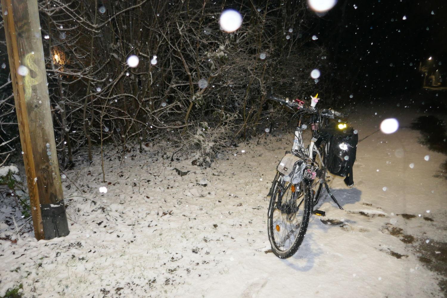 ein weisses Fahrrad mit einer schwarzen Satteltasche und einem Einhornkopf auf der Lenkstange ist auf einer Schneefläche abgestellt. Das Rad hat einen zusätzlichen Gepäckträger vorn, an dem eine kleine Weisse Tasche montiert ist und eine pinkfarbene Dashcam. Das Bild wurde bei Dunkelheit mit Blitz aufgenommen - daher sehen die Schneeflocken aus, wie große, weisse Bälle, die in der Szenerie herniederhgehen