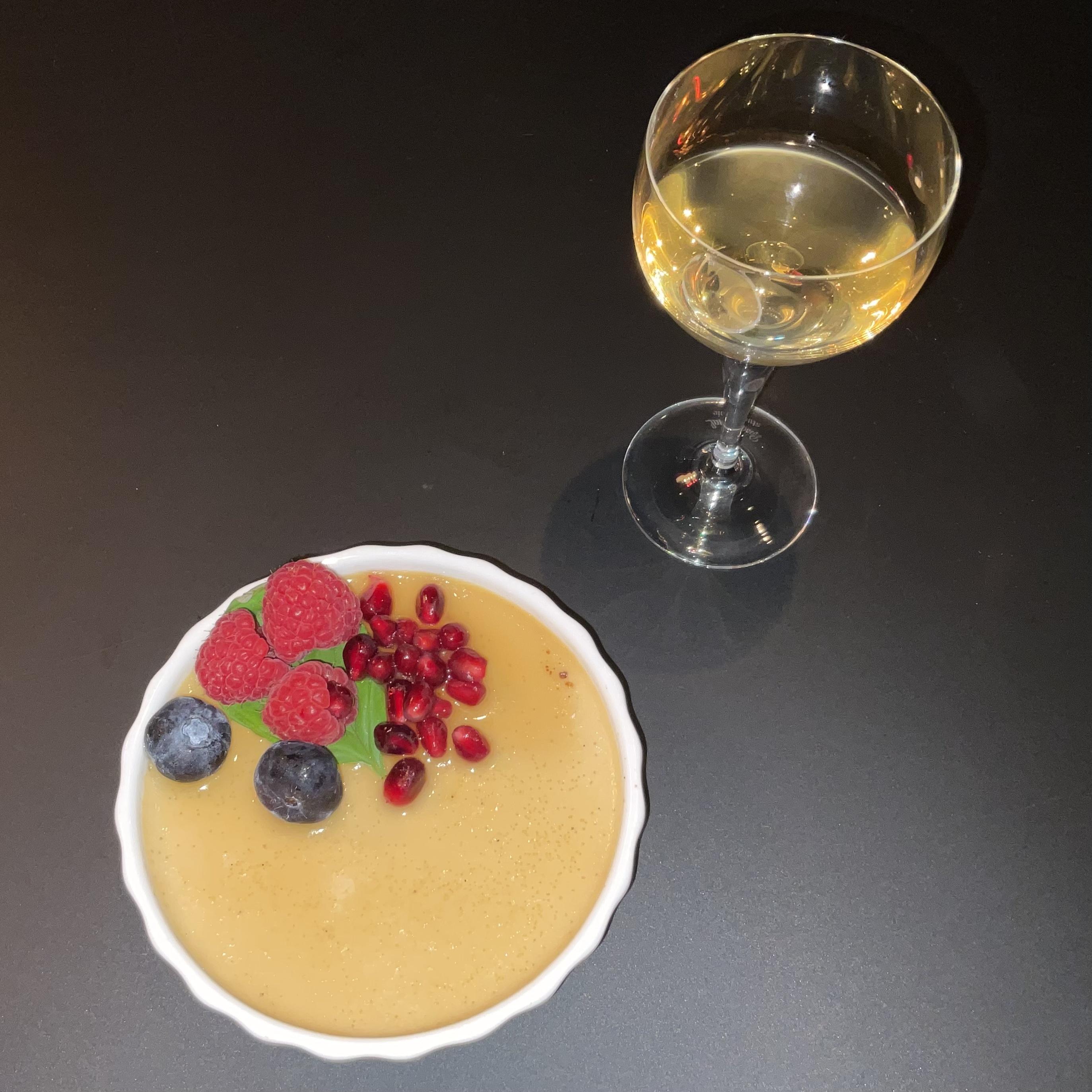 Ein weisses Tarte-Schälchen auf einer schwarzen Tischplatte, gefüllt mit Creme Brûlée, dekoriert mit einem Blatt Basilikum, ein paar Himberen, Blaubeeren und Granatapfelkernen. Dahinter rechts ein kleines Likörglas, halbgefüllt.