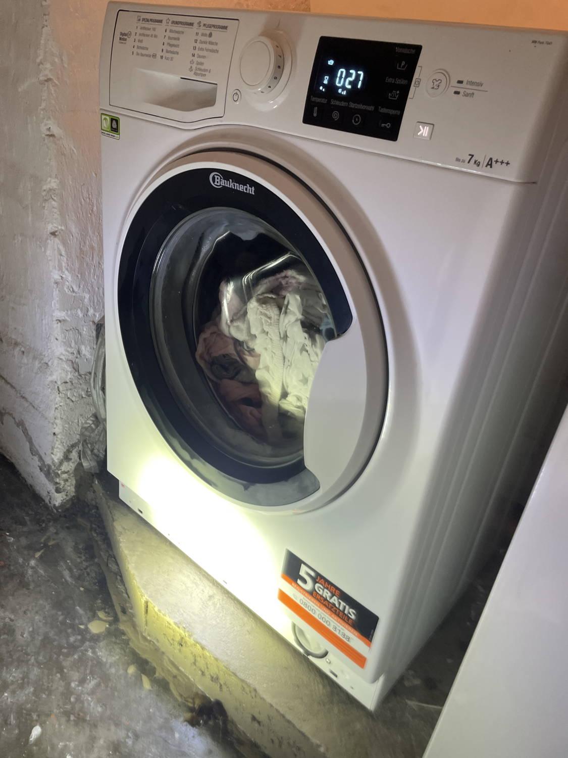 Das Bild zeigt eine Waschmaschine in einem Kellerraum. Die Maschine wäscht gerade Kinderbekleidung und auf dem Display wird 27 Minuten Restzeit angezeigt