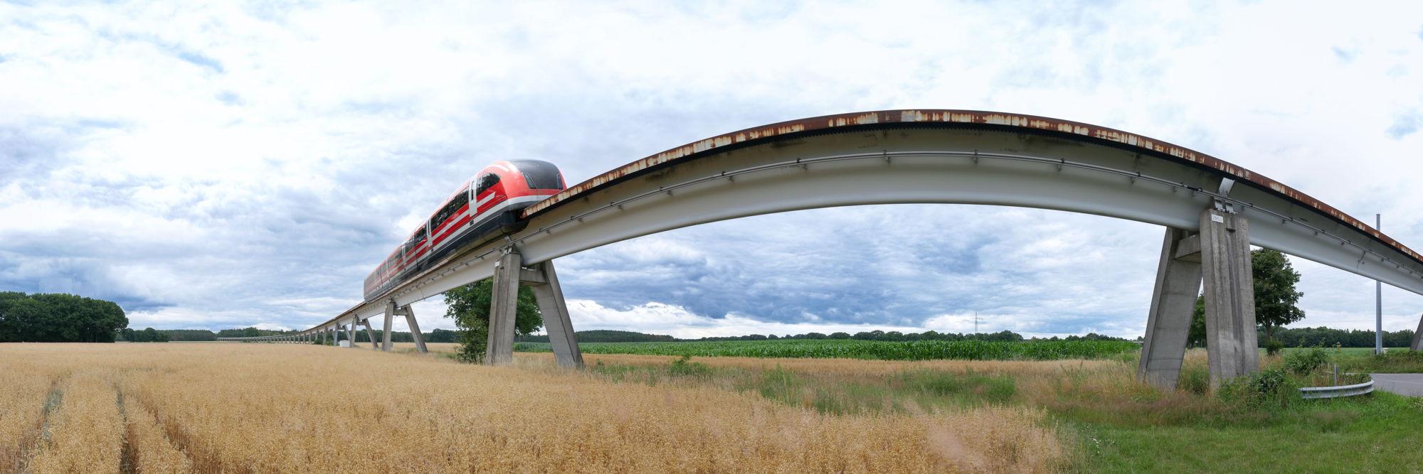 eine auf Betonpfeilern aufgeständerte Fahrspur aus Beton schlängelt sich in eleganten Bögen durch eine Landschaft mit Getreidefeldern und weiter hinten Waldbestand. Auf der Fahrtspur ist ein grau-roter Zug zu erkennen. Das Bild ist eine Fotomontage - der Zug ist auf die Fahrspur montiert worden.