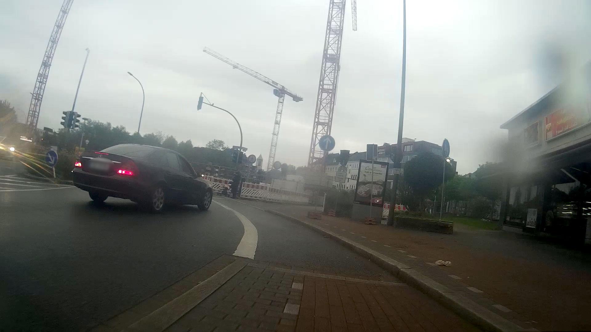 Der Radweg am Burgtor in Dortmund Richtung Norden aus der Sicht einer Fahrrad-Dashcam aufgenommen. Der Radweg wird hier auf die nach links abbiegende Straße geführt. Die schwarze Limousine steuert auf eine Einmündung auf der rechten Seite zu. Blinker ist nicht gesetzt. Die Einmündung ist mit zahlreichen Schildern zur Einfahrt gesperrt. In Hintergrund sind die Kräne einer Baustelle zu erkennen