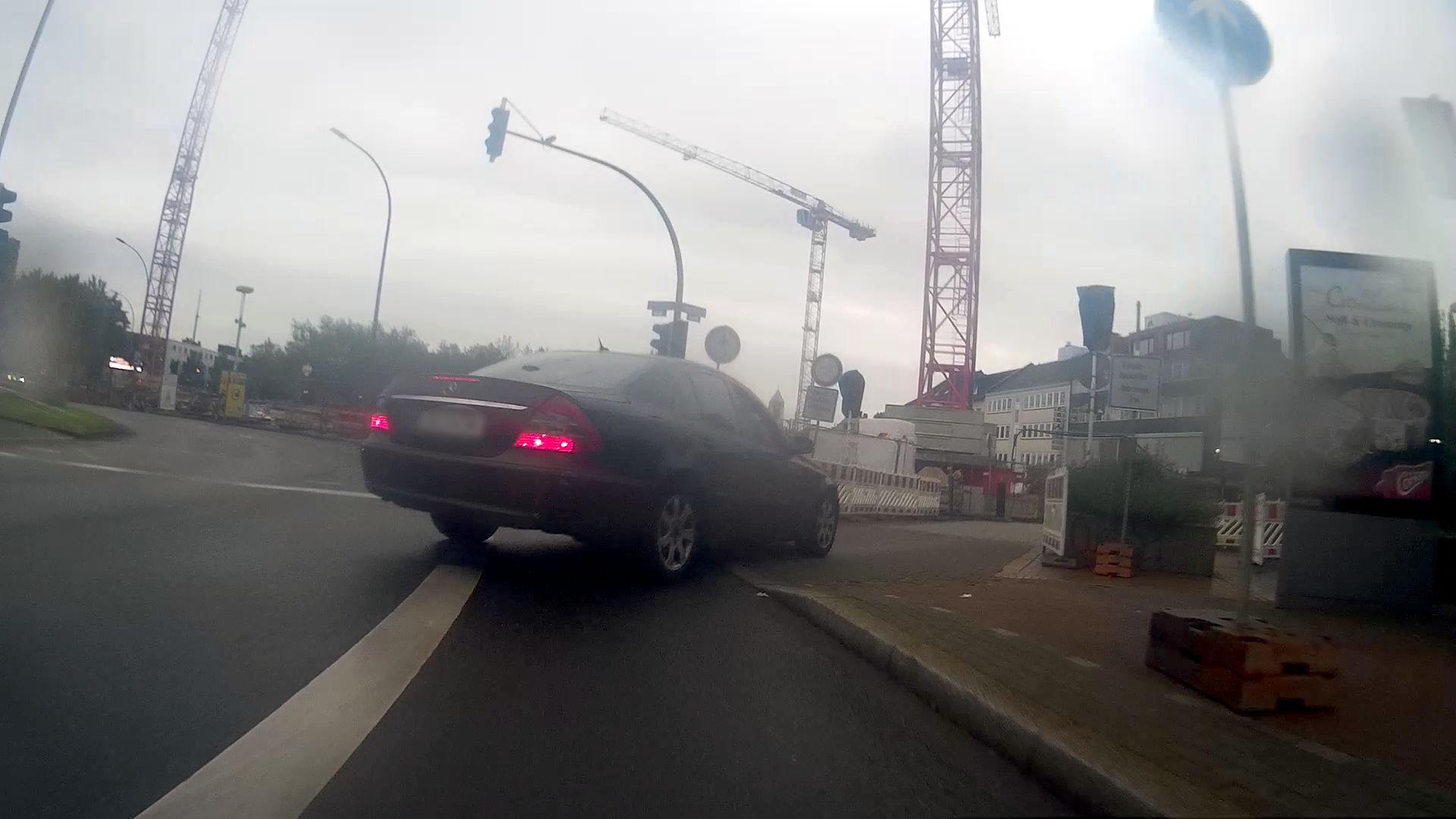 Der Radweg am Burgtor in Dortmund Richtung Norden aus der Sicht einer Fahrrad-Dashcam aufgenommen. Der Radweg läuft hier in einem Schutzstreifen auf einer nach links abbiegenden Straße. Die schwarze Limousine schneidet den Radweg auf ihrem Weg zur Einmündung auf der rechten Seite im Abstand von etwa zwei Metern zum Fahrrad. Blinker ist nicht gesetzt. Die Einmündung ist mit zahlreichen Schildern zur Einfahrt gesperrt. In Hintergrund sind die Kräne einer Baustelle zu erkennen